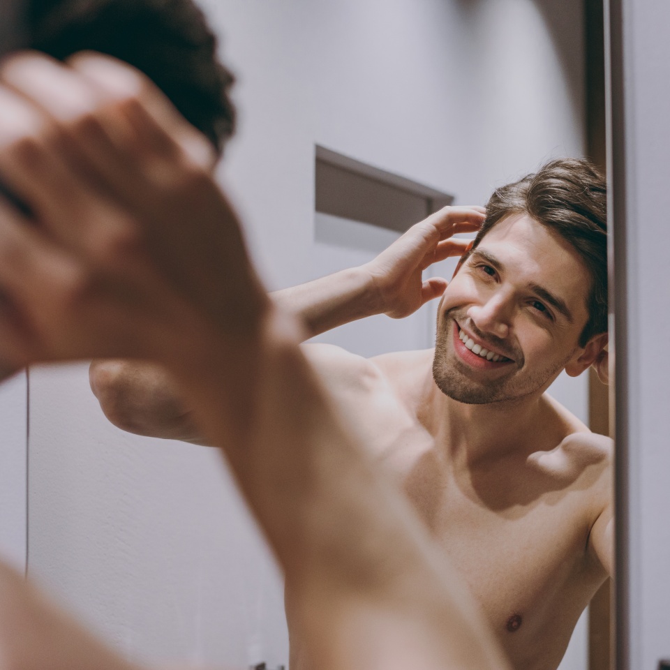 Young smiling satisfied happy attractive half naked topless caucasian brunette man 20s look at mirror touch hair indoors in bathroom at home. People healthcare daily morning routine lifestyle concept